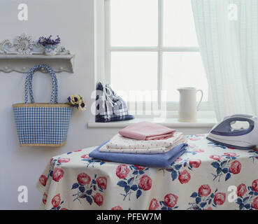 Rose patterned material draped over ironing board, folded fabrics and iron placed on top, chequered and tartan bags displayed in background, side view Stock Photo