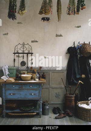 A rustic room containing kitchen utensils on a blue table, dried plants hanging from ceiling, coats on hooks on wall, wicker basket and shoes on floor Stock Photo