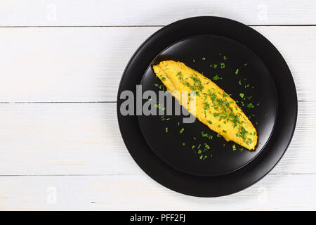 classic basic French three eggs fluffy omelette sprinkled with finely chopped parsley on black plate,  on white wooden table, view from above Stock Photo