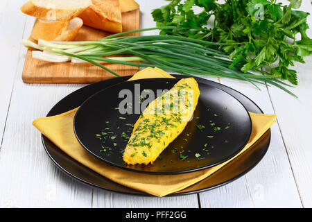 delicious French tender omelette served on black plates with yellow napkin, on white wooden table with spring onions, fresh parsley and french baguett Stock Photo