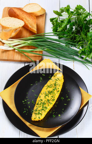 delicious French tender omelette served on black plates with yellow napkin, on white wooden table with spring onions, fresh parsley and french baguett Stock Photo