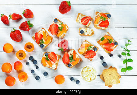 delicious strawberry apricot blueberry crisp toasts with cream cheese decorated with mint leaves with ingredients on white wooden background, easy bre Stock Photo