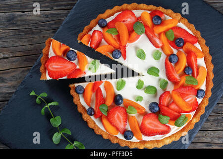 Delicious creamy cheese cake decorated with fresh ripe strawberries, apricots,  blueberries and mint  leaves with cut out one slice on slate tray on d Stock Photo