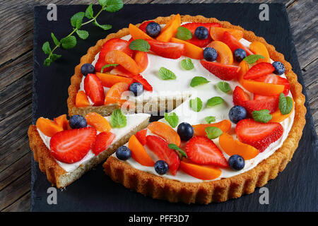 close-up of Delicious creamy cheese cake decorated with fresh ripe strawberries, apricots,  blueberries and mint  leaves with cut out one slice on sla Stock Photo