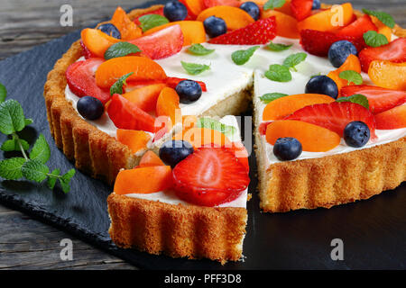 close-up of Delicious creamy cheese cake decorated with fresh ripe strawberries, apricots, blueberries and mint leaves with cut out one slice on slate Stock Photo