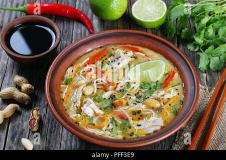 asian Chicken noodle soup sprinkled with sesame, finely chopped coriander, peanuts and decorated with a lime slices in clay bowl, soy sauce chili pepp Stock Photo