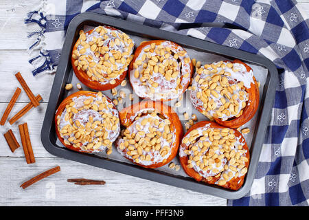 delicious freshly baked homemade cinnamon rolls topped with peanuts on baking sheet on wooden table with kitchen towel and cinnamon sticks, view from  Stock Photo