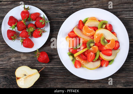 delicious fruit salad of strawberry, kiwi fruit and pear slices and mandarin on white dish on an old wooden table with strawberries on plate, view fro Stock Photo
