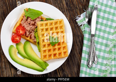 waffles sandwich with avocado, meat mushrooms and liver pate, lettuce, spinach, tomatoes on white plate on dark wooden table with fork and knife, view Stock Photo