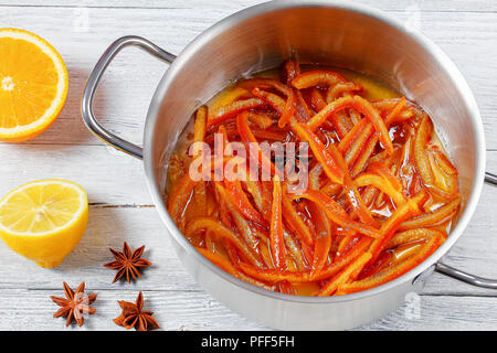 Homemade candied peels strips of orange and lemon cooked in syrup with cinnamon, nutmeg and anise star in pot on old wooden table with half of orange  Stock Photo