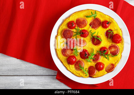 tasty omelette with sausages cut in slices, whole cherry tomatoes and fresh arugula on white platter on red table mat, view from above Stock Photo
