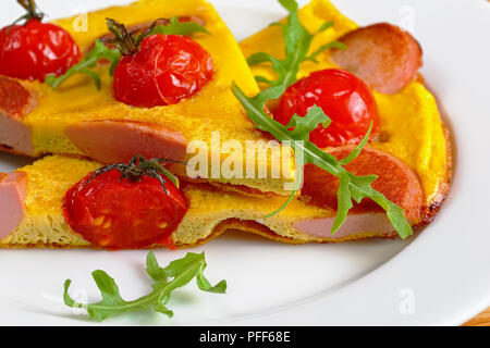 portion of tasty omelette with sausages cut in slices, whole cherry tomatoes and fresh arugula on white plate, close-up, macro Stock Photo