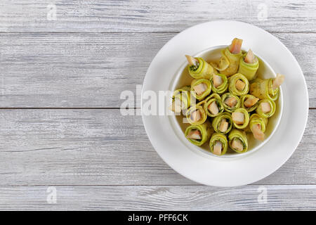 Delicious marinated in vinegar zucchini rolls filling with herring slices and dill in white bowl on wooden table, view from above Stock Photo