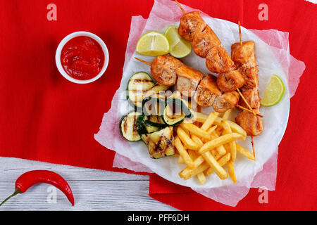 delicious chicken shish kebabs on skewers served with french fries, grilled zucchini slices and lemon slices on white plate on table with ketchup in g Stock Photo