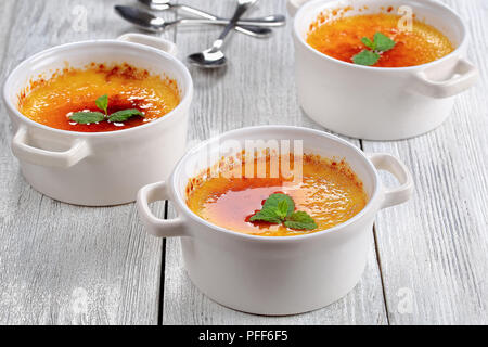tasty Creme brulee or Trinity cream with hard caramel top, decorated with mint leaves in white ceramic ramekins on table, dessert spoons on background Stock Photo