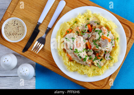 delicious Rabbit braised in sour cream with mushrooms and carrots served with mashed potatoes on white plate on wooden board with fork and knife, view Stock Photo