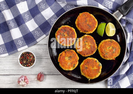 delicious fresh fried homemade fish cakes with mashed potato on skillet with lime slices, on wooden table with kitchen towel and mix of peppercorn, au Stock Photo