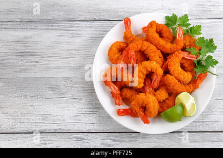 delicious deep fried breaded cheese prawn tails served with lime wedges and coriander leaves on white plate on wooden table, blank space for text left Stock Photo