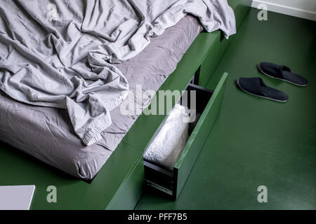 Stylish bedroom in a modern style with white wall and green floor. There is a green bed with drawers and pastel linens, black slippers on the floor. C Stock Photo