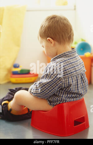 Boy toddler sitting on plastic potty with trousers down, view from behind. Stock Photo