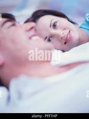 Man in white shirt and woman in blue top lying on bed, smiling, woman looking at man, side view. Stock Photo