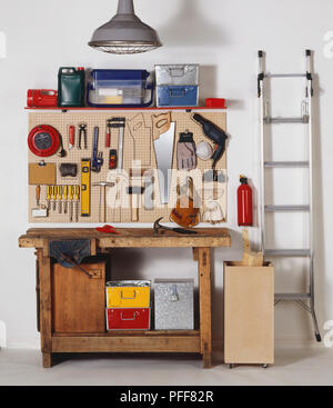 Workbench, metal boxes, work tools hanging on wooden board on wall, ladder leaning on wall,  and storage cases on wall shelf, front view. Stock Photo