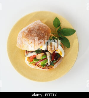Pan Bagnat, top of bread roll lifted to reveal filling of boiled egg, anchovy, tuna, red onion rings, salad and tomato, garnished with basil leaves and served on yellow plate, view from above. Stock Photo