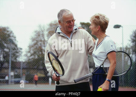 Mature man holding tennis racket, mature woman with racket under arm and tennis ball in hand, each smiling and with arm around other, on tennis court. Stock Photo