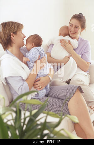 Two women sitting on sofa opposite each other, each holding a baby Stock Photo