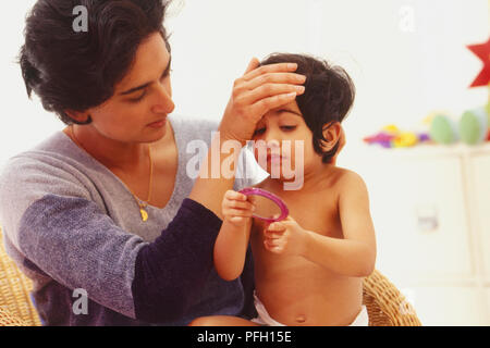 Dark haired Woman sitting in chair with girl toddler in her lap, one hand on girl's forehead. Stock Photo