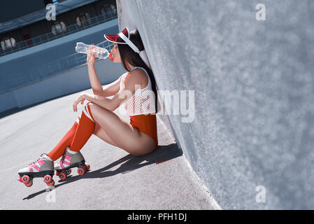 Young woman on roller skates and helmet drinking water. Stylish beautiful girl in shorts and a purple shirt rollerblading. Hipster girl resting after active time in urban skate park. Stock Photo