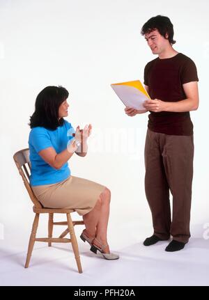 Man stood reading from a pad, woman sat on chair applauding Stock Photo