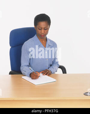 Woman in blue shirt sitting at desk and making notes, front view Stock Photo