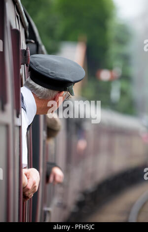 Rear view of ticket inspector (in peaked cap) leaning out of vintage carriage window as train approaches station. Taken travelling on board train. Stock Photo