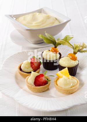 Choux pastry tartlets and chocolate cases filled with cream and garnished with strawberries and physalis, on a plate, bowl of cream nearby Stock Photo