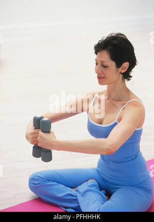 Woman sitting on mat with crossed legs, holding weights at chest height Stock Photo