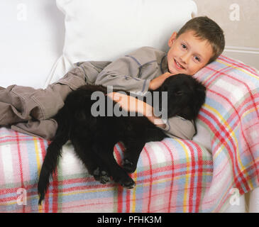 Boy lying on sofa cuddling black puppy, front view. Stock Photo