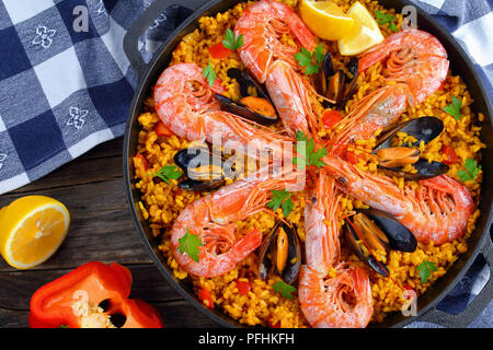 close-up of delicious seafood valencia paella with king prawns, mussels on savory creamy saffron rice with spices and lemon wedges in pan, on wooden t Stock Photo