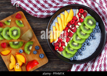 healthy breakfast- delicious chia seeds pudding with raspberry, blueberries, peach and kiwi fruit slices in black bowl on old wooden table with ingred Stock Photo