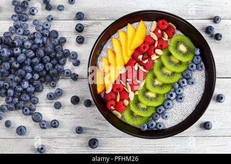 delicious chia seeds pudding with raspberry, blueberry, peach and kiwi fruit slices in black bowl on old wooden table, with  pile of blueberries on ba Stock Photo