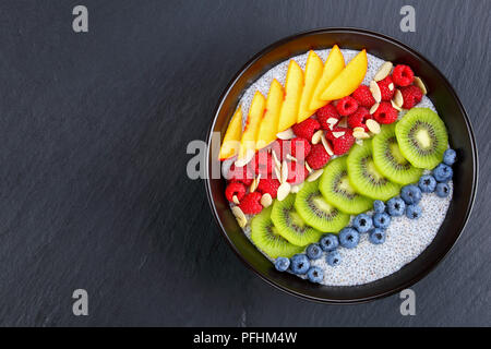 delicious chia seeds pudding with raspberry, blueberry, peach and kiwi fruit slices sprinkled with almond flakes in black bowl on dark slate tray, vie Stock Photo