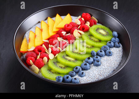 delicious chia seeds pudding with raspberry, blueberry, peach and kiwi fruit slices sprinkled with almond flakes in black bowl on dark slate tray, vie Stock Photo