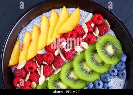 close-up of delicious chia seeds pudding with raspberry, blueberry, peach and kiwi fruit slices sprinkled with almond flakes in black bowl on stone tr Stock Photo