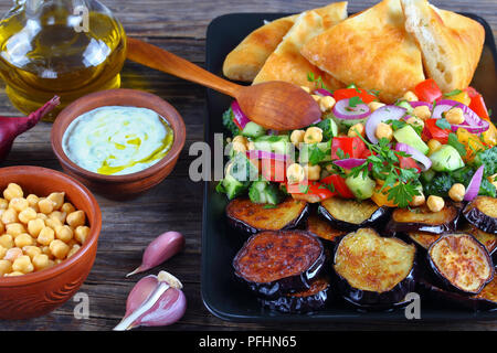 Mediterranean chickpea Salad with cucumber, red onion, tomato and fried eggplants on black dish with pita bread, tzatziki sauce and spoon on dark wood Stock Photo