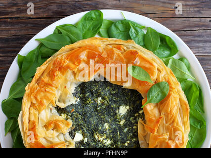delicious crusty hot greek spinach feta cheese pie or spanakopita on white platter with fresh spinach leaves, on old dark wooden table, authentic reci Stock Photo