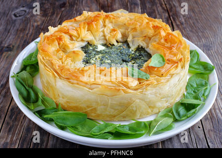 delicious golden crust hot greek spinach feta cheese pie or spanakopita on white platter with fresh spinach leaves, on old dark wooden table, classic  Stock Photo