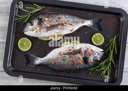 Fresh raw dorado or sea bream fish with lemon, herbs, oil, vegetables and sprinkled with spices on baking dish, on wooden worktop, top view Stock Photo