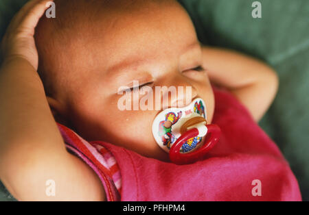 Baby girl in pink outfit sleeping with a dummy in her mouth and arms over her head, close up. Stock Photo
