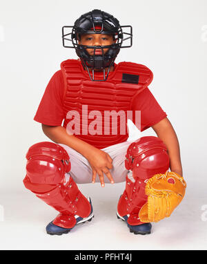 Boy dressed in baseball protective clothing, crouching down wearing face mask, chest guard, fielders mit and leg guards Stock Photo