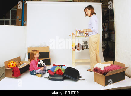 Woman next to shelf looking down at girl sitting cross-legged on floor near open suitcase and three cardboard boxes full of toys and fabrics Stock Photo